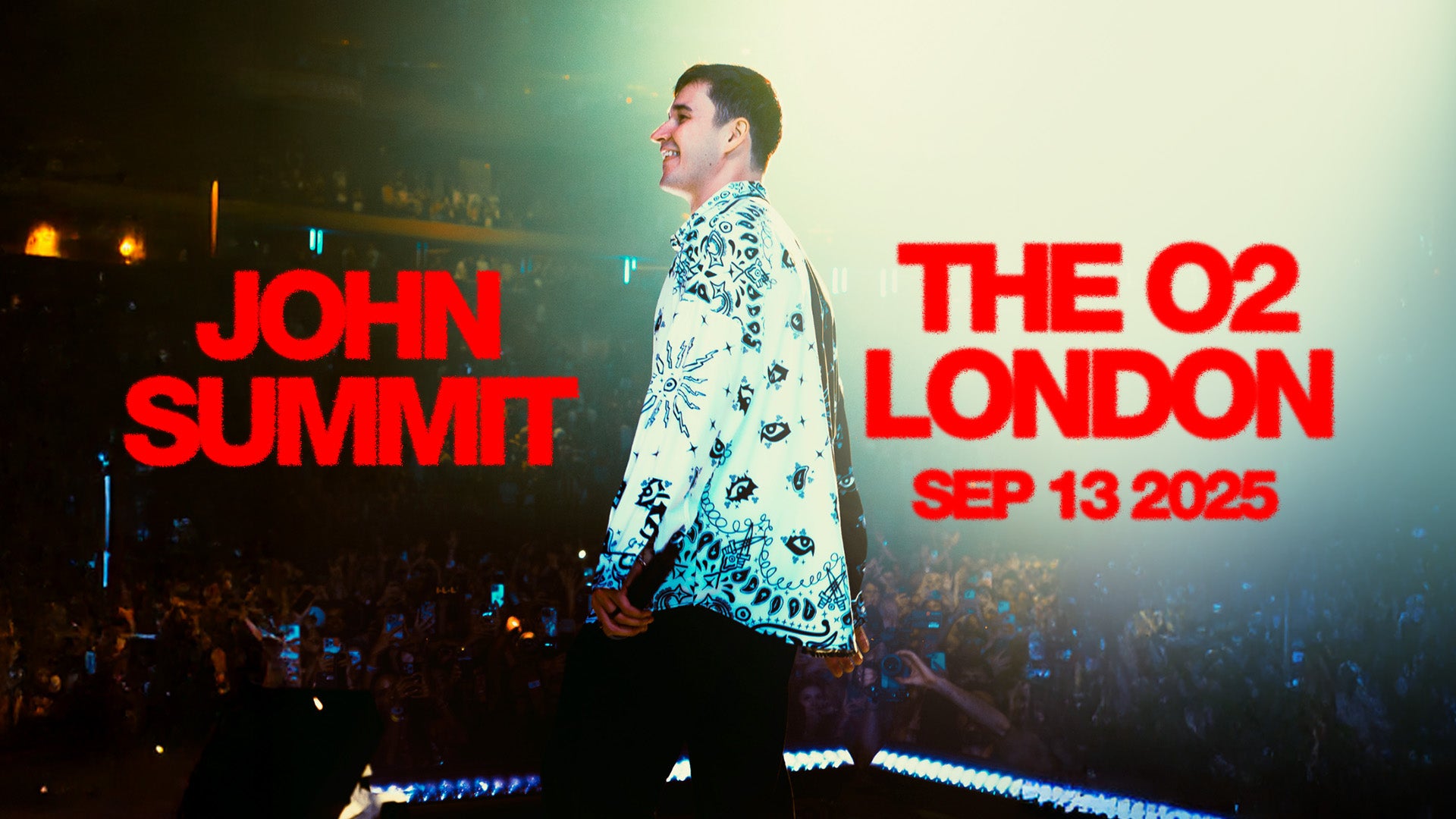 Promotional image for John Summit’s concert at The O2. He is shown on stage, smiling and facing the crowd, wearing a white shirt with black patterns. The stage and audience are lit with colourful lights.