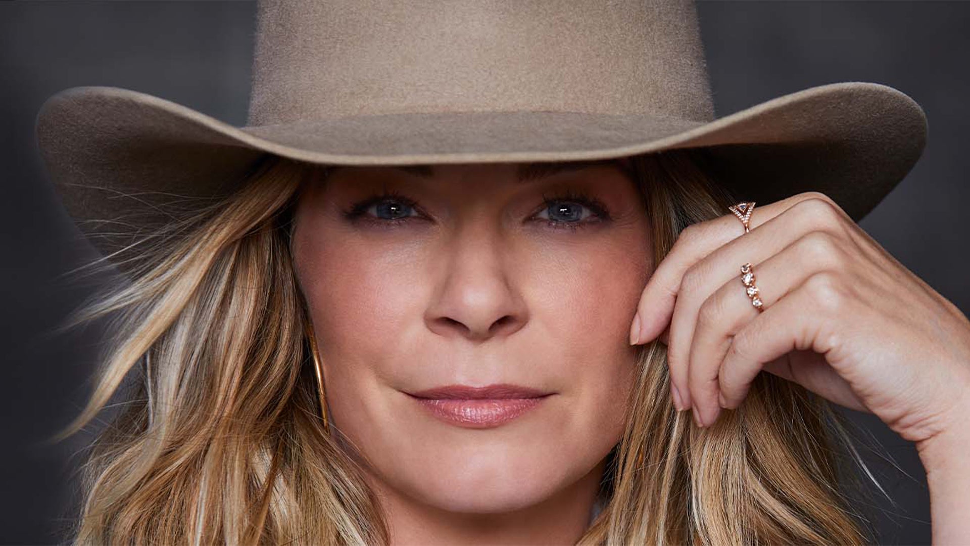 Close-up portrait of singer Leanne Rimes who has blonde hair wearing a beige hat and a ring, resting her chin on her hand, with a neutral gray background.