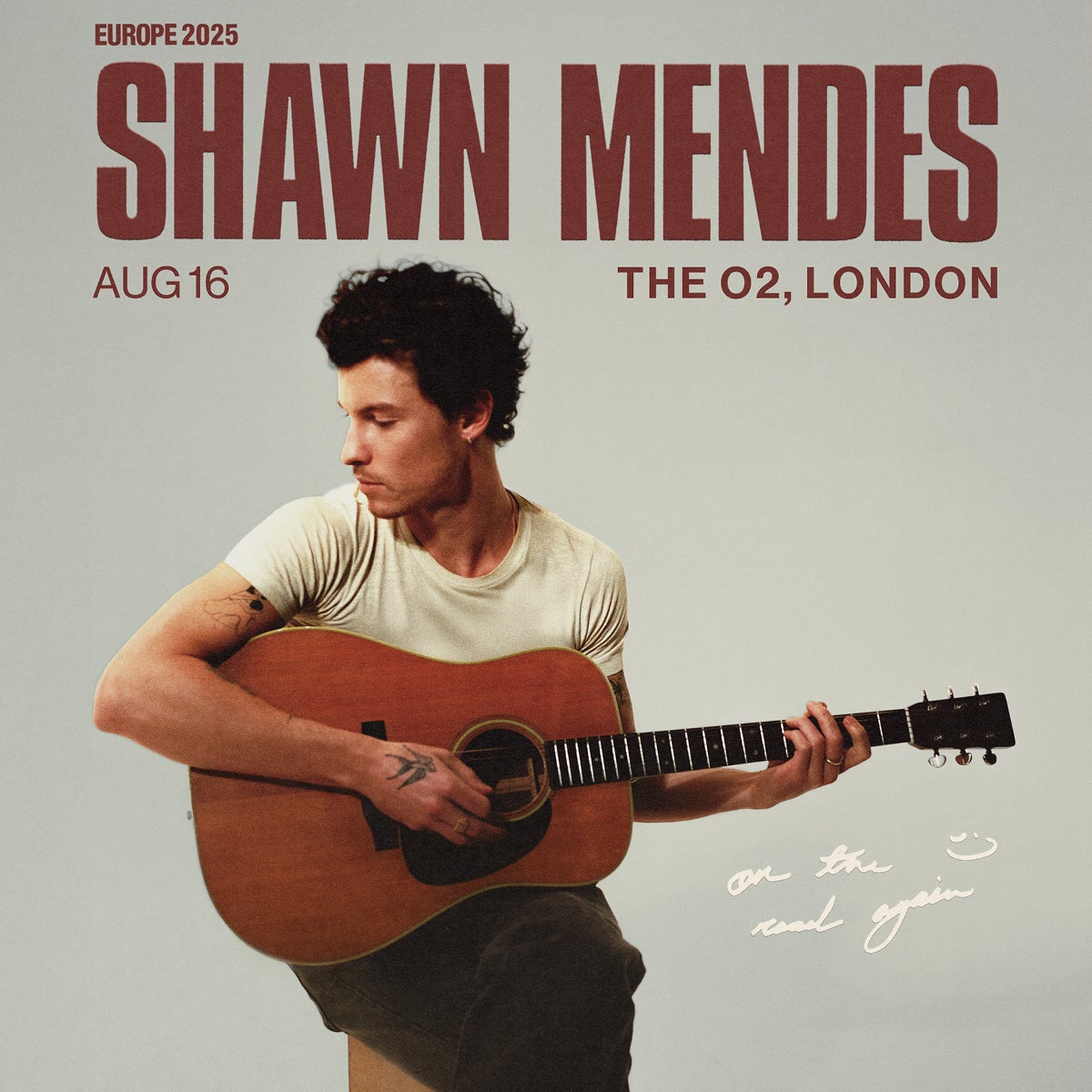 A promotional poster for Shawn Mendes’ Europe 2025 tour. The poster features a photo of Shawn Mendes seated, playing an acoustic guitar, looking down and to the side. The text reads “SHAWN MENDES, AUG 16, THE O2, LONDON, Europe 2025.” There’s a handwritten note near the bottom of the image that says “on the road again” with a smiley face doodle. Shawn is wearing a simple t-shirt and has visible tattoos on his arms.