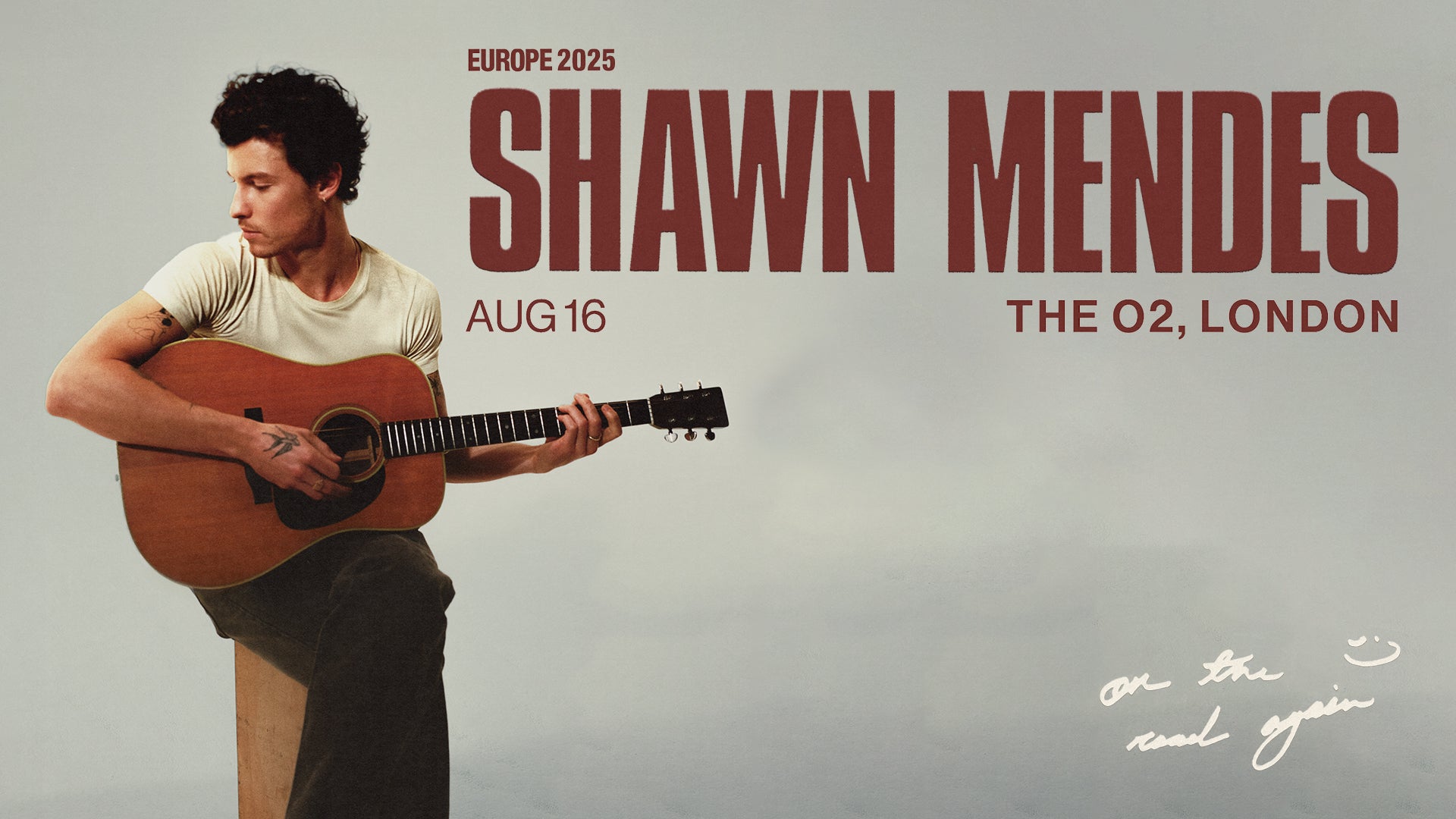 A promotional poster for Shawn Mendes’ Europe 2025 tour. The poster features a photo of Shawn Mendes seated, playing an acoustic guitar, looking down and to the side. The text reads “SHAWN MENDES, AUG 16, THE O2, LONDON, Europe 2025.” There’s a handwritten note near the bottom of the image that says “on the road again” with a smiley face doodle. Shawn is wearing a simple t-shirt and has visible tattoos on his arms.